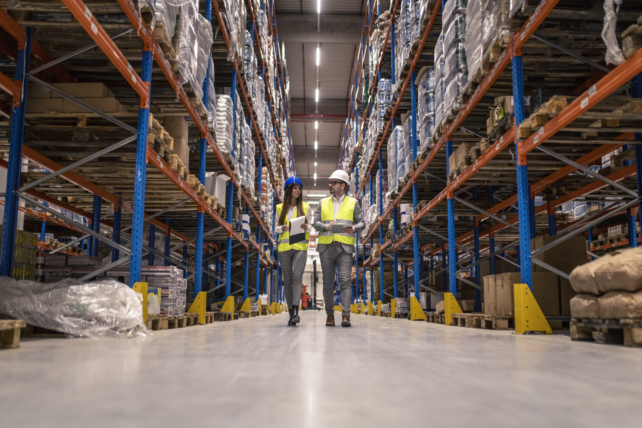 Workers with hardhats and reflective jackets walking through big warehouse aisle checking goods condition.