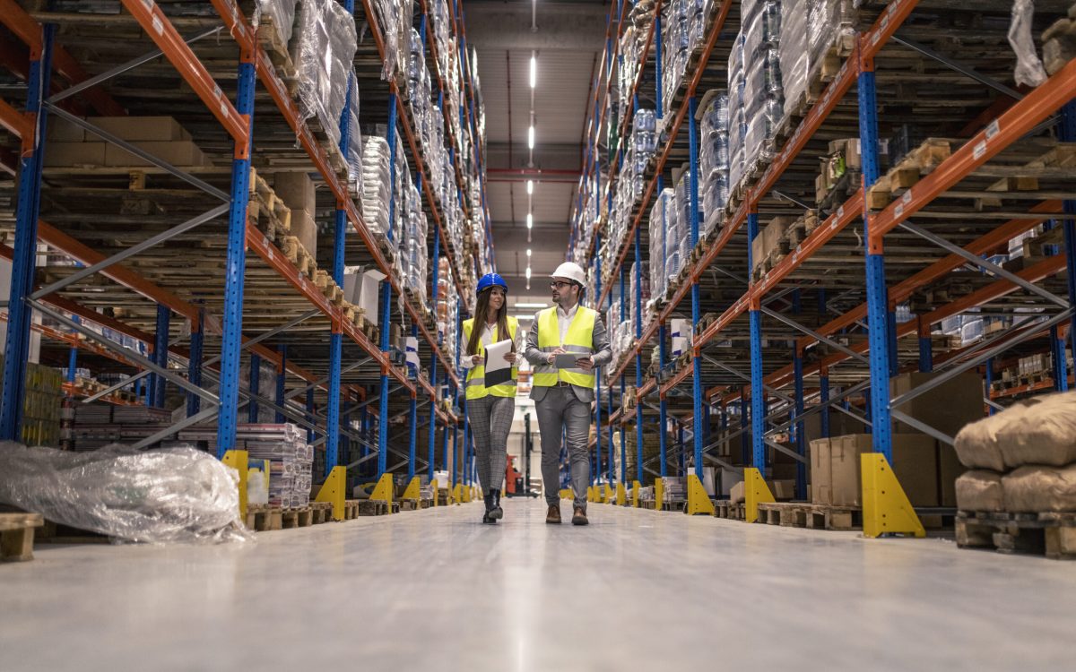 Workers with hardhats and reflective jackets walking through big warehouse aisle checking goods condition.